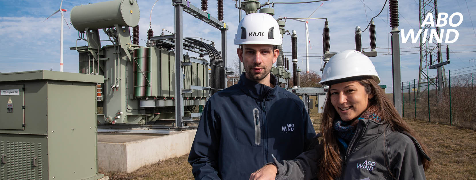 Mann und Frau mit Bauhelmen stehen vor Umspannstation und Windkraftanlagen