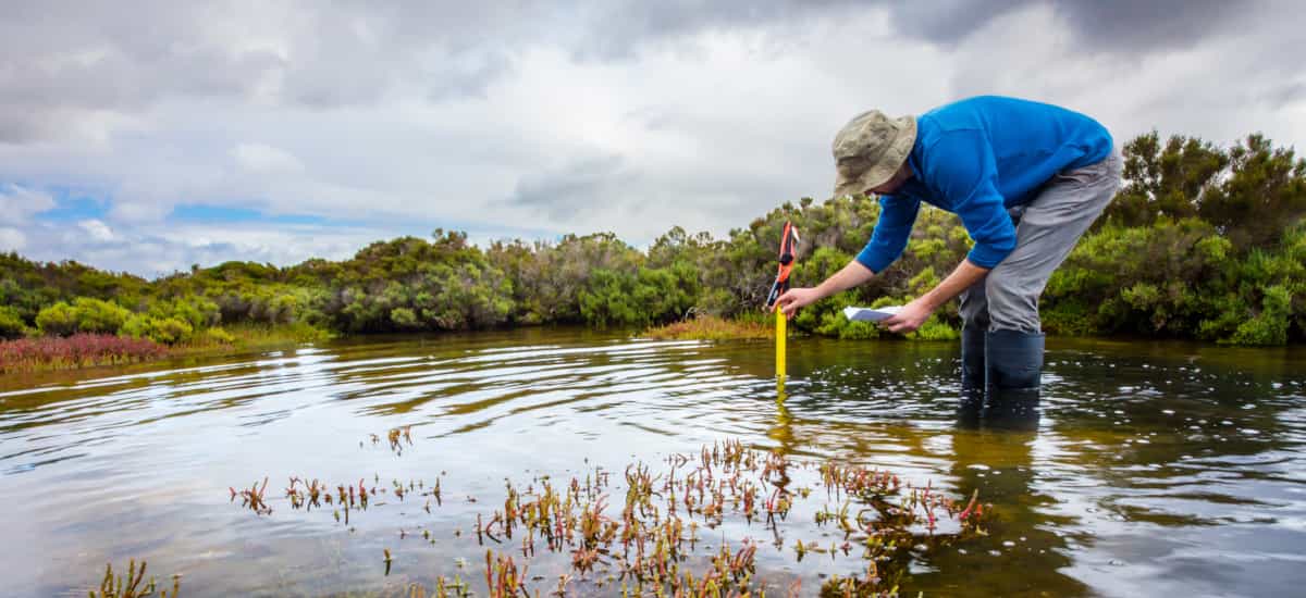 Mann steht gebückt in einem flachen See und misst die Wassertiefe. Der Himmel ist bewölkt und im Hintergrund am Ufer stehen Büsche und Bäume. - Bild von IzzetNoyan (Adobe Stock) Mann steht gebückt in einem flachen See und misst die Wassertiefe. Der Himmel ist bewölkt und im Hintergrund am Ufer stehen Büsche und Bäume.