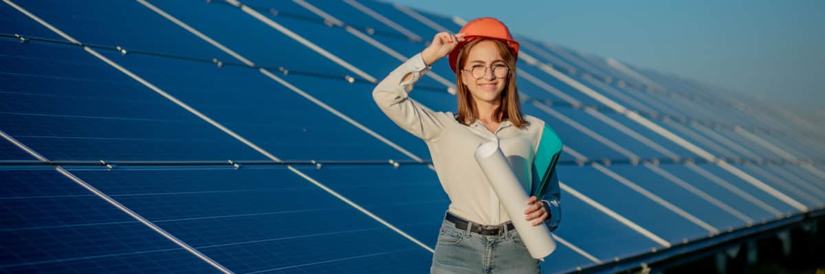 Frau steht mit Bauhelm und Planrolle vor Freiflächen-Photovoltaikanlage