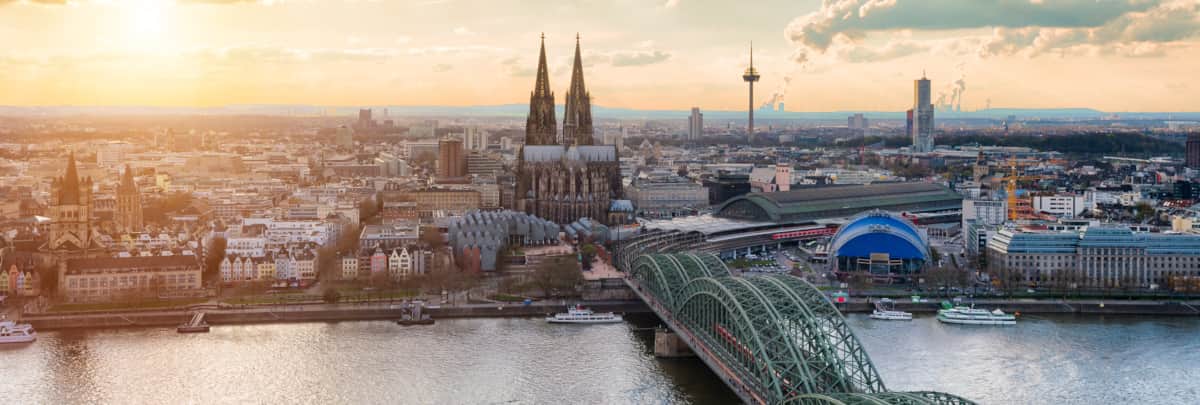 Blick auf Köln. Zentral im Bild der Dom und Bahnhof. Im Vordergrund der Rhein und die Eisenbahnbrücke (Hohenzollernbrücke). Im Hintergrund geht langsam die Sonne unter.