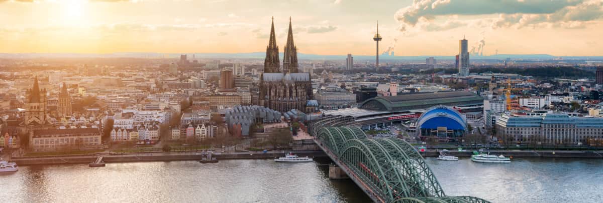 Blick auf Köln. Zentral im Bild der Dom und Bahnhof. Im Vordergrund der Rhein und die Eisenbahnbrücke (Hohenzollernbrücke). Im Hintergrund geht langsam die Sonne unter. - Bild von Simon (Adobe Stock) Blick auf Köln. Zentral im Bild der Dom und Bahnhof. Im Vordergrund der Rhein und die Eisenbahnbrücke (Hohenzollernbrücke). Im Hintergrund geht langsam die Sonne unter.