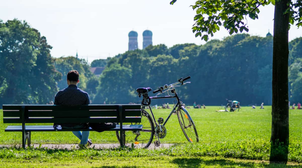 Mann sitzt entspannt auf einer Parkbank (von hinten gesehen), daneben steht ein Fahrrad. Dahinter eine große Rasenfläche mit Menschen, im Hintergrund Bäume und dahinter hervorragenden Türmen der Münchener Frauenkirche.